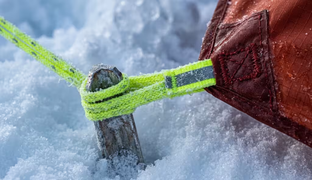 Close-up of a 4 season backpacking tent reinforced anchor point with a reflective guyline secured to a snow picket for maximum stability in winter camping.